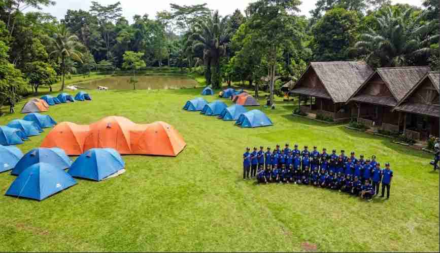 Pemandangan udara area perkemahan hijau luas di Telaga Cikeas dengan banyak tenda biru dan oranye, sekelompok besar orang berbaju biru sedang berfoto bersama di dekat pondok kayu tradisional di depan danau.